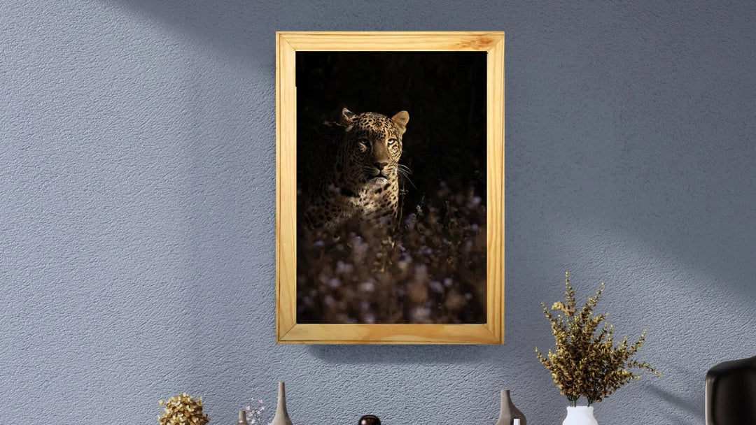 A framed photographic print of a leopard in a dark natural setting, hung on a textured blue-gray wall above decorative vases and dried plants.