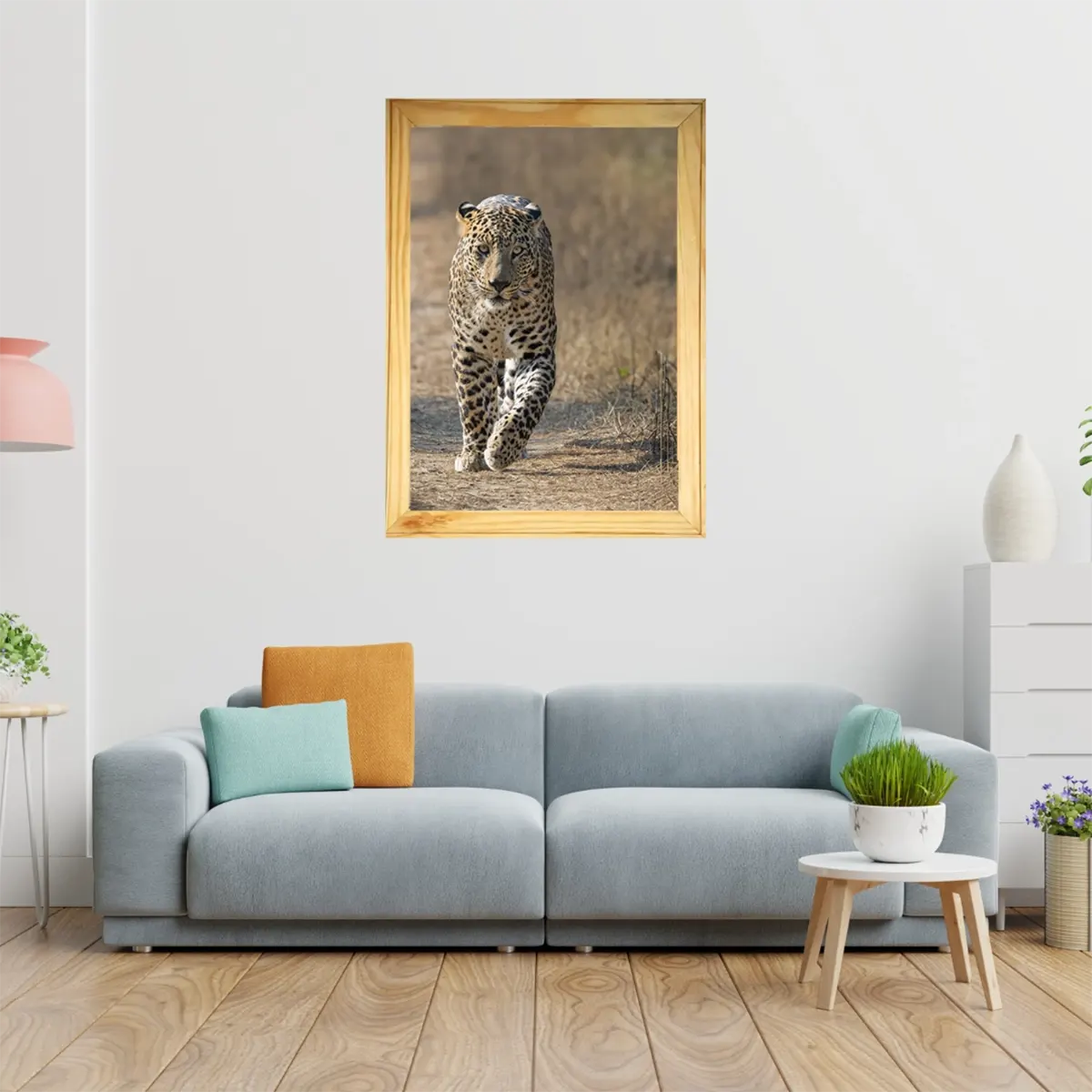 A framed vertical art print of a walking leopard in a golden-brown savanna, displayed on a white wall above a modern light blue sofa in a styled living room.