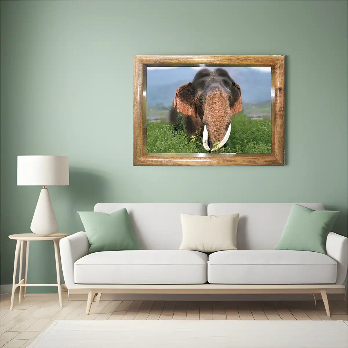 A framed wall art print of an elephant in a lush green field, displayed on a sage green wall above a modern white sofa with mint and cream pillows in a styled living room.